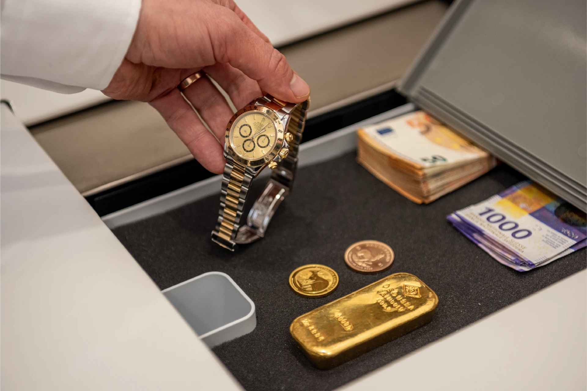 Hand taking a luxury watch out of a safe deposit box with a gold bar, gold coins and euro banknotes
